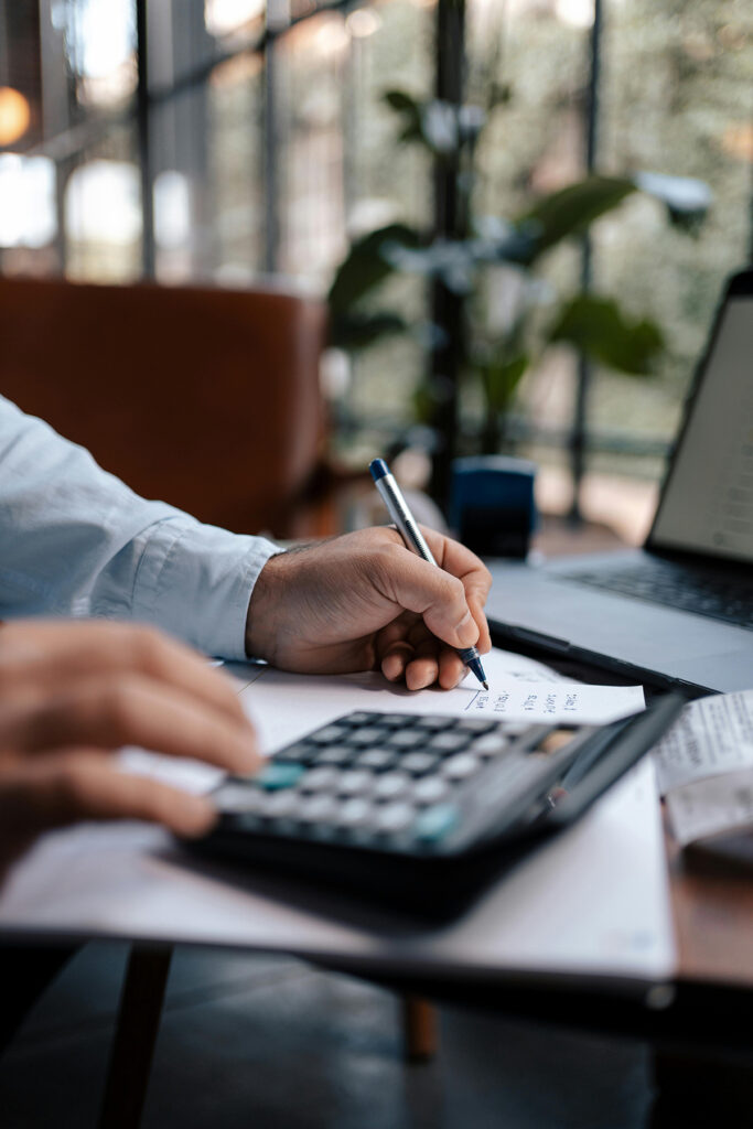 A person's hands holding a pen and writing while also working a calculator by a laptop